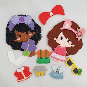 May include: A collection of felt doll accessories, including two doll faces with dark hair, various outfits, a red bow, and yellow sunglasses. The outfits include a purple top with a green purse, orange overalls, a red dress, and a blue dress.