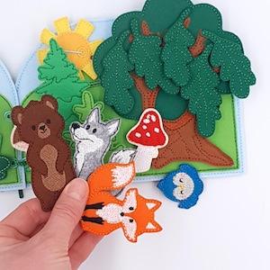 May include: A felt forest scene featuring a brown bear, grey wolf, orange fox, red mushroom, and blue owl. The scene includes a large green tree, a yellow sun, and a green and blue landscape. The felt pieces are hand-stitched.