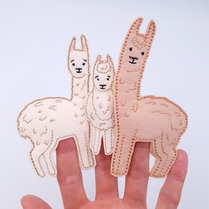 May include: Three felt llama finger puppets in cream and peach colors. The puppets have embroidered details, including eyes, mouths, and decorative stitching. The llamas are in different sizes, suggesting a family theme.