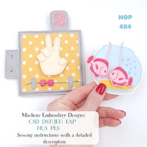 May include: A handmade felt educational toy featuring a yellow polka dot background, a hand gesture, and the number 2. The image also shows two pink fish in a blue water scene. The text "HOP 4X4" is visible.