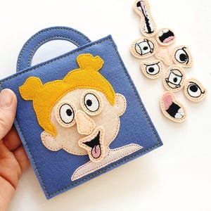 Puede incluir: Un libro de actividades de fieltro azul con una cara cosida y cabello amarillo. El libro tiene un asa e incluye varios rasgos faciales de fieltro, como ojos y bocas, para un juego interactivo.