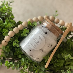 May include: A glass jar filled with white bath salts with a cork lid and a wooden spoon. The jar has a white label that says "Foot Soak Deluxe Salts" and "Handmade in CA Cedarbrook Essentials".