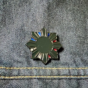 May include: A black metal pin with a sunburst design. The sunburst has eight points and is outlined in silver. The centre of the sunburst is black and has a red, white, and blue design.