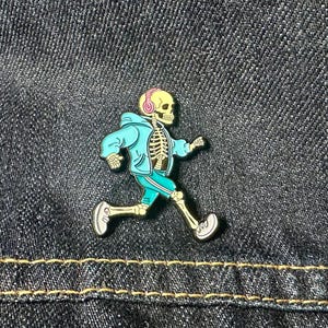 Może przedstawiać: Szkielet w niebieskim swetrze i słuchawkach biegnie z determinacją. Szkielet ma na sobie białe buty sportowe i ma żółtą czaszkę.