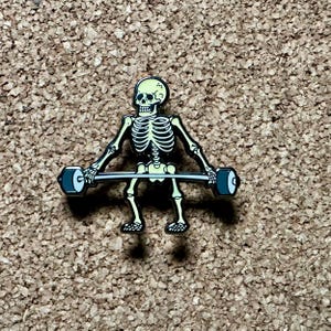 Könnte beinhalten: Ein Skelett, das eine Langhantel mit schwarzen Gewichten hebt. Das Skelett ist gelb und schwarz und trägt ein schwarzes Stirnband. Die Langhantel ist silber.