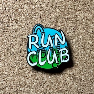May include: A blue, green, and white enamel pin with the words "RUN CLUB" and a stylized image of a running shoe.