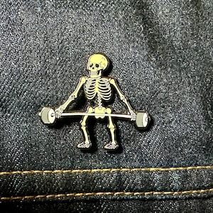 May include: A yellow skeleton lifting a barbell with black weights. The skeleton is wearing a gold-colored pin.