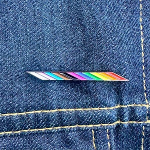 May include: A rainbow pride pin with a silver metal base and a diagonal stripe design. The pin features a variety of colors, including red, orange, yellow, green, blue, indigo, and violet.