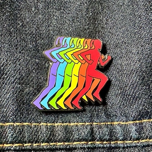 May include: A rainbow-coloured enamel pin depicting a running figure. The design shows a sequence of runners in shades of purple, blue, green, yellow, orange, and red. The pin is set against a denim background.