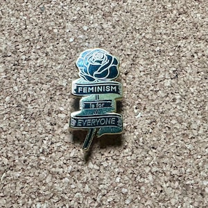 May include: Gold-colored pin with a black rose and a banner that reads "Feminism is for everyone".