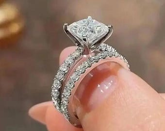 Könnte beinhalten: Ein silberner Verlobungsring mit einem quadratisch geschliffenen Diamanten und einem mit Diamanten besetzten Band. Der Ring wird hochgehalten, um sein Design und Glitzern hervorzuheben. Ein zweites Band ist ebenfalls sichtbar.