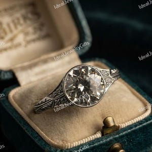 May include: A silver-toned ring featuring a large, round, faceted center stone set in a bezel with milgrain detailing. The band is crafted with intricate, engraved geometric patterns and small accent stones along the shoulders of the ring.