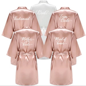 Op de afbeelding: Een verzameling satijnen badjassen in verschillende kleuren, waaronder blush roze en wit. Elke badjas heeft een andere titel in witte letters op de rug: "Bridesmaid", "Bride", "Team Bride", "Mother of the Bride" en "Maid of Honor".