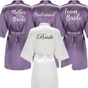 Könnte beinhalten: Satz von vier Satin-Morgenmänteln in Lila und Weiß mit Text auf der Rückseite. Die Morgenmäntel sagen "Mutter der Braut", "Braut", "Brautjungfer" und "Team Braut".