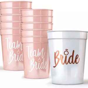May include: A set of pink plastic cups with the text "Team Bride" in white lettering and a gold ring icon. There is also a white plastic cup with the text "Bride" in gold lettering and a gold ring icon.
