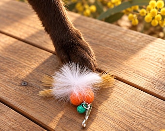 May include: A cat toy featuring two small orange felt balls, a tuft of white feathers, and a small green metal bell attached to a silver metal safety pin. The toy is positioned on a wooden surface near a dark brown animal paw.