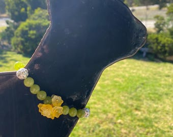 May include: A beaded bracelet with green and yellow beads, and flower-shaped charms. The bracelet is displayed on a black cat-shaped silhouette against a backdrop of green grass and trees.