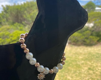 May include: A beaded bracelet with white faux pearls and gold-colored beads. A gold-colored charm in the shape of a flower hangs from the bracelet. The bracelet is displayed on a black cat-shaped stand against a backdrop of greenery and a blue sky.