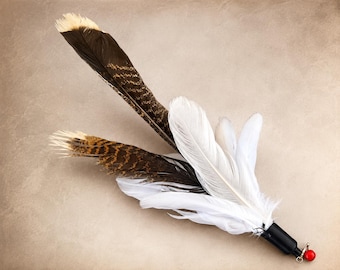 May include: A cat toy featuring a cluster of natural brown and black striped feathers mixed with solid white feathers. The feathers are attached to a black cylindrical base with a small silver metal ring and a single bright red spherical bead.
