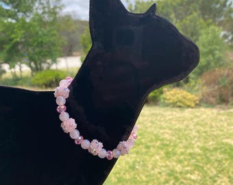 May include: A pink beaded bracelet with various sized beads and floral accents is displayed on a black cat-shaped stand. The bracelet features a mix of light pink and clear beads, creating a delicate and feminine accessory.