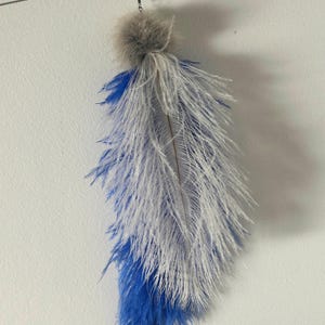 May include: A hanging decorative ornament featuring a cluster of long, wispy white and vibrant blue ostrich feathers. The feathers are gathered at the top with a soft, fluffy grey pom-pom and attached to a thin metal wire hook for suspension.