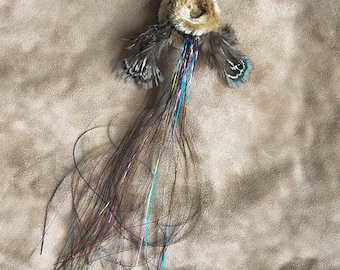 May include: A decorative keychain featuring a circular tan faux fur pom-pom, two dark brown and teal patterned feathers, and a long, flowing tassel made of thin, multicolored metallic strands attached to a silver metal lobster clasp.