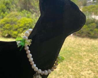 May include: A black cat-shaped jewelry display with a pearl necklace. The necklace features white and pink pearls, green floral accents, and a yellow pendant. The background shows a green landscape and a blue sky.