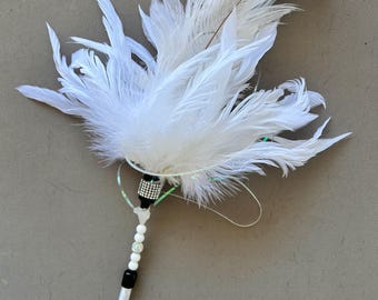 May include: A white feather wand featuring a cluster of soft white plumes attached to a handle. The handle is decorated with a black and silver rhinestone-encrusted band, followed by a series of white beads and a single sparkling rhinestone bead.