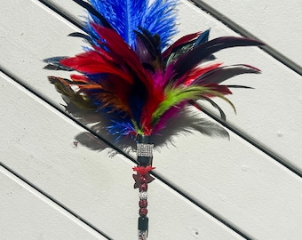 May include: A decorative wand featuring a cluster of vibrant blue, red, green, and black feathers at the top. The handle is black with a silver rhinestone band, a small red butterfly accent, and a clear lower section filled with colorful glitter.