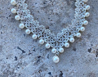 May include: A silver-toned necklace featuring a V-shaped floral pattern encrusted with clear crystals and accented with round white pearls. The design includes a central dangling pearl drop and additional pearls spaced along the metal frame.