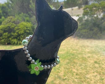 May include: A black metal cat silhouette with a beaded necklace. The necklace features gray beads, green accents, and a green and pink flower. The cat is positioned in front of a window with a view of green foliage and a cloudy sky.