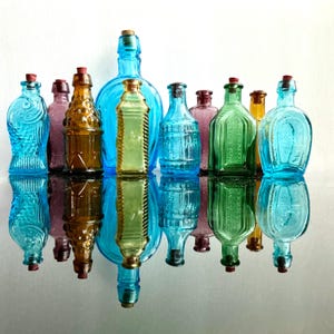 May include: A collection of colorful glass bottles with cork stoppers. The bottles are in various shapes and sizes, including a fish-shaped bottle and a bottle with a textured design. The bottles are in shades of blue, green, brown, and pink.