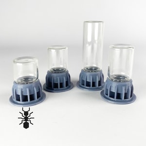 May include: Four clear glass vials of different heights, each sitting on a grey, 3D-printed base. The bases have a circular design with vertical supports. The vials are empty and cylindrical, suitable for small items or samples.