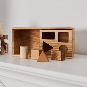 Könnte beinhalten: Eine hellfarbene Holzbox zum Formensortieren mit verschiedenen geometrischen Blöcken, ein Igel-Plüschtier mit braunem Karo-Outfit und Brille sowie zwei hölzerne Vogel-Wanddekorationen, die über der weißen Oberfläche an der Wand befestigt sind.