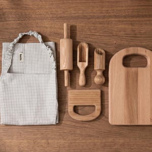 Puede incluir: Un delantal de cuadros vichy gris claro y blanco con una etiqueta Kavka, un rodillo de madera, una pala de madera, un mortero de madera, un raspador de masa de madera y una tabla de cortar de madera con la marca Kavka grabada abajo a la derecha.