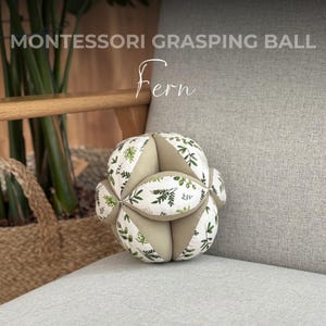 May include: A Montessori grasping ball with a white background and green leaf pattern. The ball is labeled "Fern".
