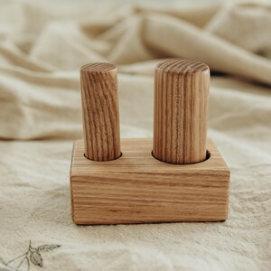 May include: A wooden toy featuring two cylindrical pegs set in a rectangular base. The pegs and base are made of light-colored wood, showcasing the natural grain. The toy is designed for early childhood development.