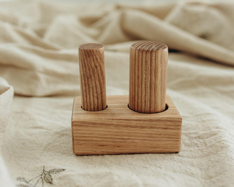 May include: A wooden toy featuring two cylindrical pegs set in a rectangular base. The pegs and base are made of light-colored wood, showcasing the natural grain. The toy is designed for early childhood development.