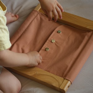 Könnte beinhalten: Ein Spielzeug aus pfirsichfarbenem Stoff mit Holzrahmen und drei goldfarbenen Knöpfen. Das Spielzeug wird von einem Kind bedient. Der Rahmen besteht aus hellem Holz. Der Stoff ist über den Rahmen gespannt.