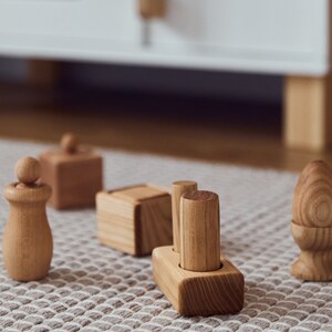May include: A collection of wooden toys, including a figure with a ball on top, a cube with a lid, a block with two cylinders, and an egg-shaped piece. The toys are made of light-colored wood and are arranged on a patterned rug.