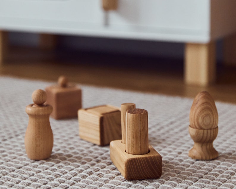 May include: A collection of wooden toys, including a figure with a ball on top, a cube with a lid, a block with two cylinders, and an egg-shaped piece. The toys are made of light-colored wood and are arranged on a patterned rug.