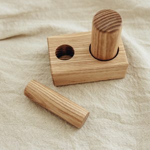 May include: A wooden toy set featuring a rectangular block with two holes and two cylindrical pieces. The wood has a natural grain and a light brown color. One cylinder is inserted into a hole, and the other is separate.