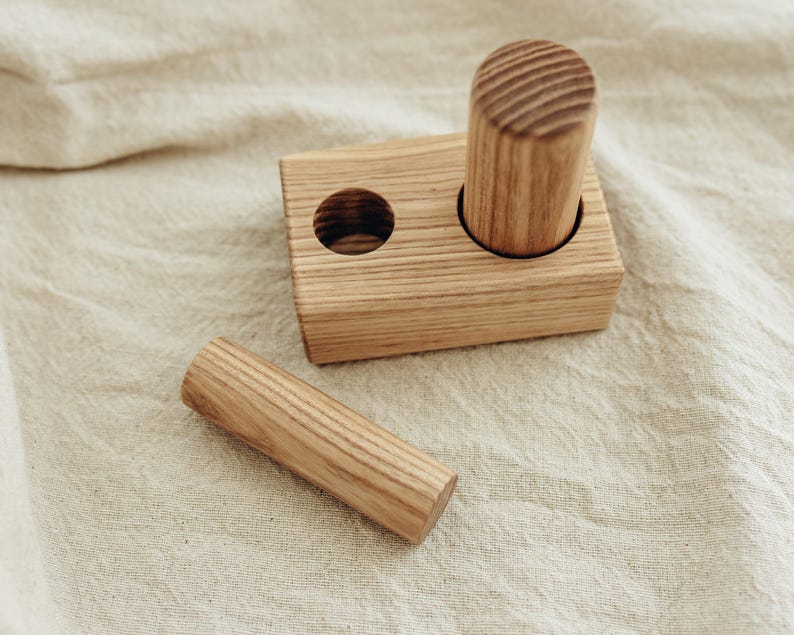 May include: A wooden toy set featuring a rectangular block with two holes and two cylindrical pieces. The wood has a natural grain and a light brown color. One cylinder is inserted into a hole, and the other is separate.