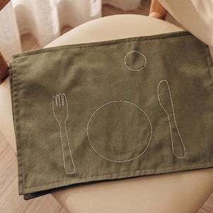Könnte beinhalten: Ein rechteckiges, olivgrünes Platzset mit weißen Stickereien, die eine Gabel auf der linken Seite, einen Teller in der Mitte, ein Messer auf der rechten Seite und eine kleine kreisförmige Tassen- oder Glasform in der oberen rechten Ecke zeigen.