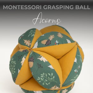 May include: A yellow and green fabric ball with an acorn and leaf pattern. The ball is made of soft fabric and is designed for grasping and sensory play. Montessori Grasping Ball Acorns.
