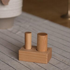 May include: A wooden block with two cylindrical pegs. The block is rectangular with two circular holes. One peg is taller than the other. The wood has a light brown color and a visible grain.