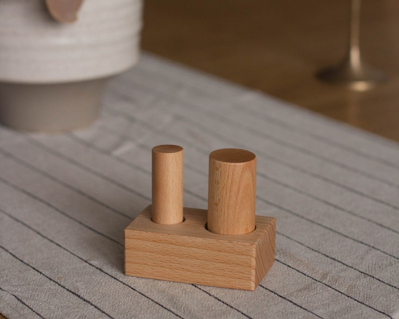 May include: A wooden block with two cylindrical pegs. The block is rectangular with two circular holes. One peg is taller than the other. The wood has a light brown color and a visible grain.