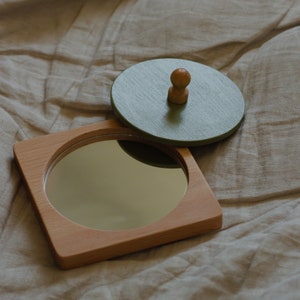 Puede incluir: Una caja de madera con un espejo redondo en el interior y una tapa verde con un pomo de madera.