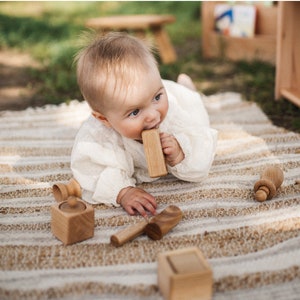 May include: A baby in a white outfit lies on a striped rug, chewing on a wooden block. Several other wooden toys, including a small box, a spoon, and a bottle-shaped toy, are scattered around. The scene is set outdoors with natural light.