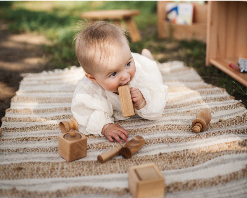 May include: A baby in a white outfit lies on a striped rug, chewing on a wooden block. Several other wooden toys, including a small box, a spoon, and a bottle-shaped toy, are scattered around. The scene is set outdoors with natural light.